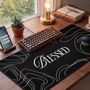 May include: A black desk mat with the word "BLESSED" in white script. White abstract lines decorate the mat. A wooden keyboard, mouse, and phone are on the desk. A small succulent plant in a terracotta pot sits nearby.