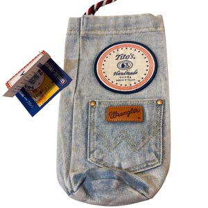 May include: A light blue denim bottle bag featuring a round Tito's Handmade Vodka patch, a Wrangler pocket with a brown leather logo tag, and a red, white, and blue braided drawstring cord. A product tag with text and a QR code is attached.