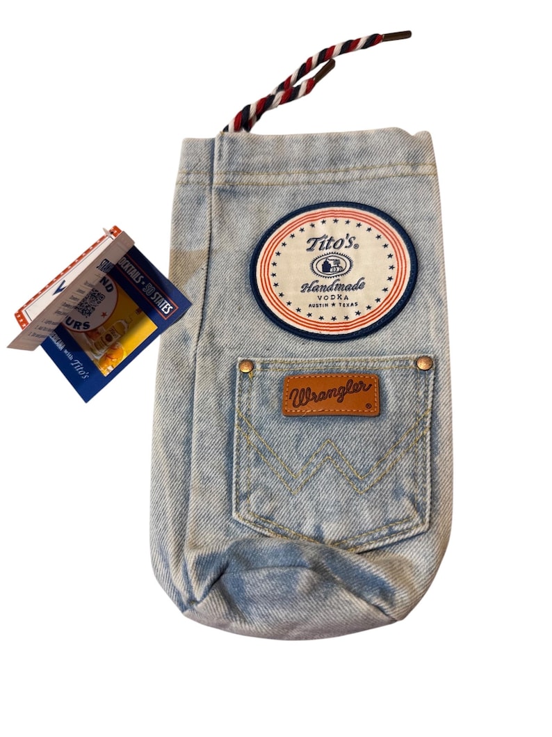 May include: A light blue denim bottle bag featuring a round Tito's Handmade Vodka patch, a Wrangler pocket with a brown leather logo tag, and a red, white, and blue braided drawstring cord. A product tag with text and a QR code is attached.
