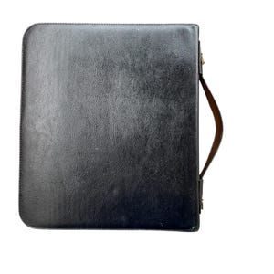 May include: A black leather portfolio with a textured surface. It features a brown leather handle attached to the side with a metal clasp. The portfolio is rectangular with rounded corners and a zipper closure.