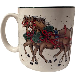 Puede incluir: Una taza de cerámica color crema con un borde verde oscuro, que presenta una ilustración gráfica de dos caballos marrones con bridas rojas y coronas verdes con cintas rojas. El fondo de la taza tiene un sutil patrón de motas.