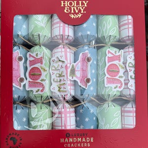 Puede incluir: Una caja de seis petardos navideños de lujo hechos a mano. Los petardos son en tonos de azul, verde y rosa, con pegatinas decorativas que dicen "JOY" y "MERRY". La caja es roja con las palabras "HOLLY & IVY" en la parte superior.