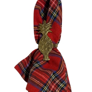 May include: A red tartan napkin is held by a gold-coloured pineapple napkin ring. The napkin has a pattern of red, black, yellow, and blue lines. The pineapple ring is detailed with textured leaves and a ring to hold the napkin.