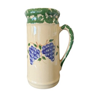May include: A tall, cream-colored ceramic pitcher with a green rim and handle. The pitcher is decorated with blue grape clusters and green leaves. The handle and rim have a textured, painted design.