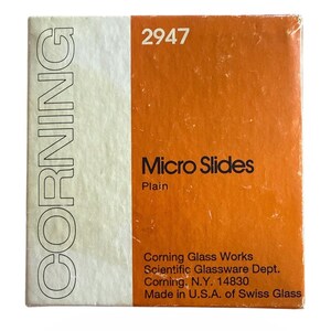 May include: A rectangular box of Corning Micro Slides, model 2947. The packaging features a white vertical strip on the left with the word CORNING printed vertically in black, and an orange background on the right with the text Micro Slides, Plain.