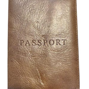 May include: A brown leather passport holder with the word "PASSPORT" embossed on the front. The cover has a textured, worn appearance. The passport holder is rectangular and appears to be closed.