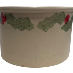 Puede incluir: Un cuenco de cerámica beige con un borde ancho. El cuenco está decorado con hojas verdes de acebo y bayas rojas alrededor de la parte superior. El cuenco tiene una superficie ligeramente texturizada y una forma redondeada.