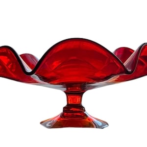 May include: A red glass bowl with a ruffled edge and a pedestal base. The bowl is made of clear glass with a red tint.
