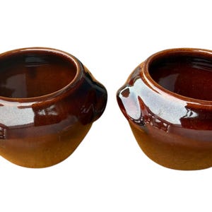 May include: Two brown ceramic pots with a glossy finish. The pots have a rounded shape with a wide opening and a small handle on the side. The interior of the pots is a darker shade of brown.