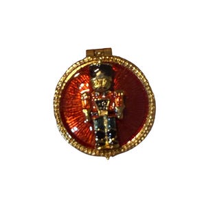 May include: A round gold-tone metal brooch featuring a central raised figure of a soldier in a red and black uniform. The background is a vibrant red enamel with a sunburst pattern, and the outer edge has a textured, beaded gold-tone border.