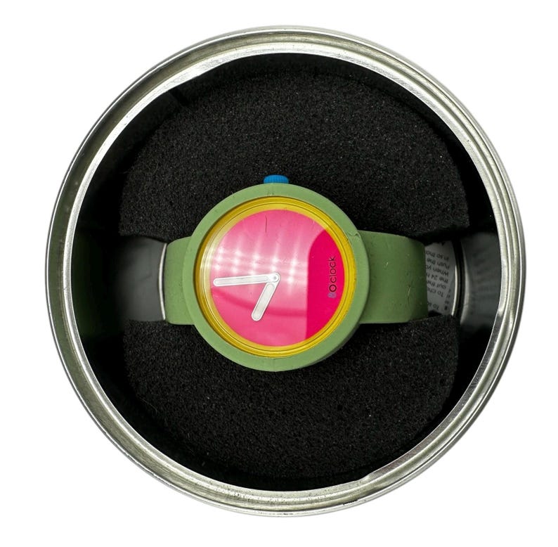 Pu&ograve; includere: Un orologio da polso verde e giallo con un quadrante rosa e lancette bianche. L'orologio &egrave; in una scatola rotonda d'argento con imbottitura in schiuma nera.