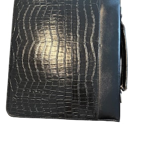May include: Black leather portfolio with a crocodile skin texture. The portfolio has a handle and a zipper closure. The overall design is sleek and professional, suitable for business or personal use.