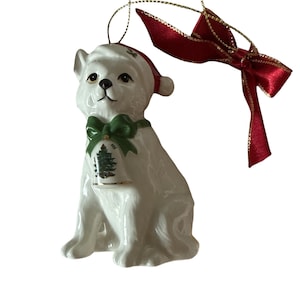 Op de afbeelding: Een witte keramische hondenkerstversiering met een rode kerstmuts en een groene strik. De hond heeft een bel om zijn nek met een kerstboomontwerp. Een rood lint met gouden rand is bevestigd.