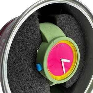 Pu&ograve; includere: Un orologio da polso verde con un quadrante rosa acceso e numeri gialli. L'orologio &egrave; in una scatola rotonda argentata con imbottitura in schiuma nera.