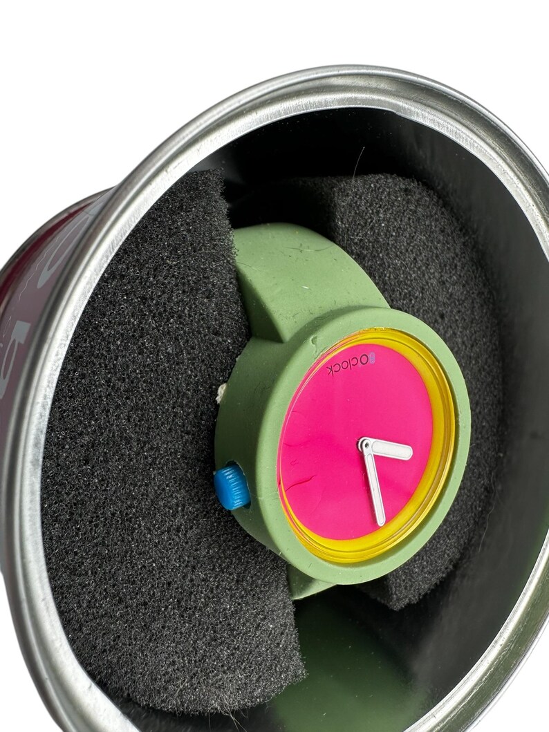 Pu&ograve; includere: Un orologio da polso verde con un quadrante rosa acceso e numeri gialli. L'orologio &egrave; in una scatola rotonda argentata con imbottitura in schiuma nera.