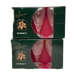 Puede incluir: Dos cajas de vasos Luminarc Epicure. Cada caja contiene tres vasos de vino de vidrio transparente con un diseño de cruz grabado. Las cajas son verdes con letras doradas y una imagen de nueces.