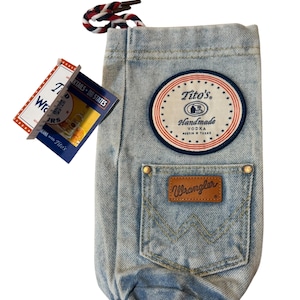 May include: A light blue denim bottle bag featuring a round patch with the text Tito's Handmade Vodka Austin Texas, a front pocket with a brown leather Wrangler label, and a red, white, and blue braided drawstring cord at the top opening.