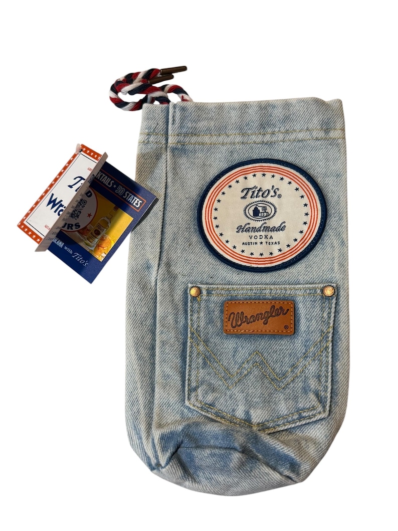 May include: A light blue denim bottle bag featuring a round patch with the text Tito's Handmade Vodka Austin Texas, a front pocket with a brown leather Wrangler label, and a red, white, and blue braided drawstring cord at the top opening.