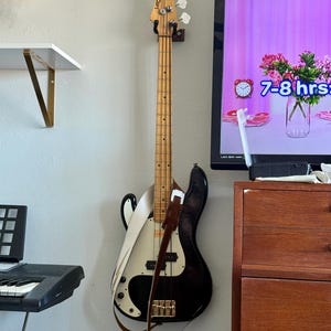 May include: A black and white electric bass guitar with a brown leather strap hanging on a wall. The bass has a wooden neck and headstock with silver tuning pegs. The body is black with a white pickguard.