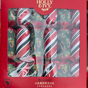 Puede incluir: Una caja roja que contiene seis petardos navideños. Los petardos alternan entre rayas diagonales rojas y blancas y un fondo verde con diseños de bastones de caramelo y acebo. La caja dice "HOLLY & IVY" y "6 LUXURY HANDMADE CRACKERS."
