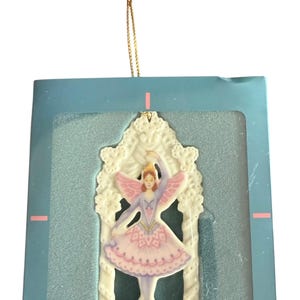 May include: A Lenox porcelain Christmas ornament featuring a ballerina in a pink tutu and wings, framed by a white decorative arch. The ornament is displayed in a teal box with a gold string for hanging.