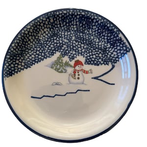 May include: A white ceramic plate with a blue and white speckled border. The plate features a winter scene with a snowman wearing a red hat and scarf.