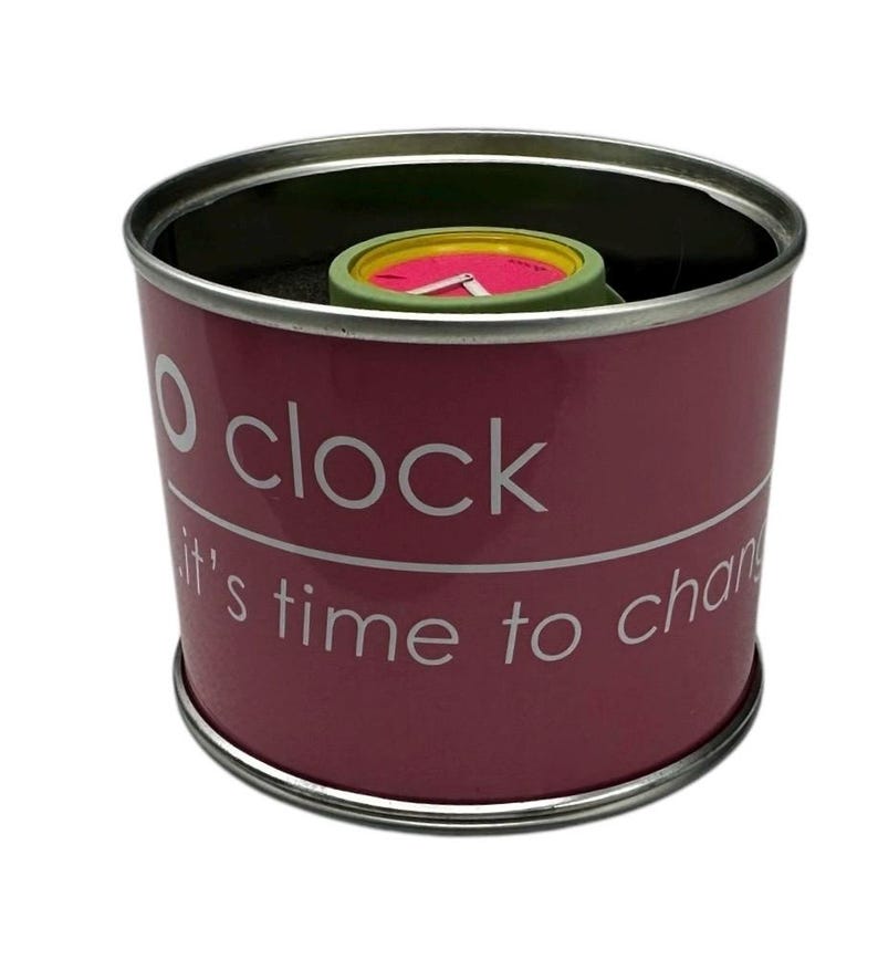 Pu&ograve; includere: Una scatola cilindrica rosa con un coperchio argentato. La scatola ha il testo "O clock it's time to change" stampato su di essa. All'interno della scatola c'&egrave; un orologio verde e rosa.
