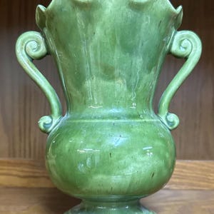 May include: Green ceramic vase with a ruffled rim and two curved handles. The vase has a bulbous body and a pedestal base. The green glaze has a mottled appearance, adding to its vintage charm. A decorative piece for displaying flowers or as a standalone accent.