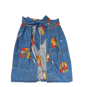 May include: Blue denim skirt with patchwork details. The skirt features a tie waist and colorful fabric patches in various patterns. The skirt has pockets and is made of denim material.