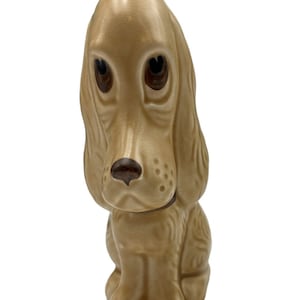 May include: A ceramic figurine of a brown dog with floppy ears and sad eyes. The dog is sitting with its head tilted down.