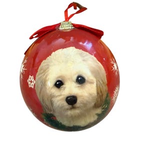 May include: Red Christmas ornament featuring a portrait of a fluffy, light-colored dog with dark eyes and nose. The ornament has a red ribbon and gold cap. White snowflakes are scattered around the dog's portrait.