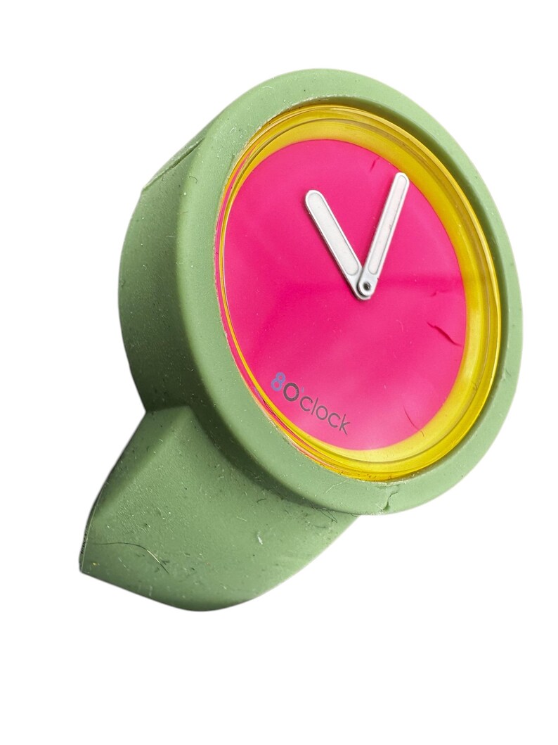 Pu&ograve; includere: Un orologio da polso in silicone verde con un quadrante rosa acceso e dettagli gialli. Il quadrante dell'orologio ha lancette bianche e il testo "8 o'clock" stampato in basso.