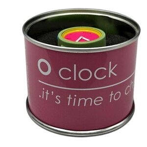 Pu&ograve; includere: Una scatola cilindrica rosa con un coperchio argentato e il testo "O clock . it's time to change" stampato sul lato. La scatola contiene un orologio da polso colorato con un quadrante verde, giallo e rosa.