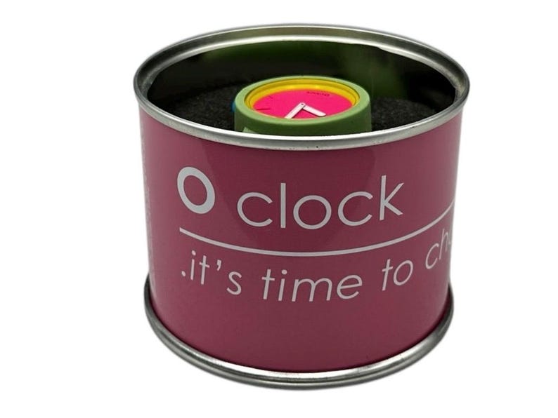 Pu&ograve; includere: Una scatola cilindrica rosa con un coperchio argentato e il testo "O clock . it's time to change" stampato sul lato. La scatola contiene un orologio da polso colorato con un quadrante verde, giallo e rosa.