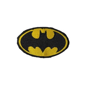 May include: An oval-shaped embroidered patch featuring a black bat silhouette centered on a vibrant yellow background, enclosed by a thick black border. The design displays the iconic winged creature emblem with sharp points and a distinct yellow outline.