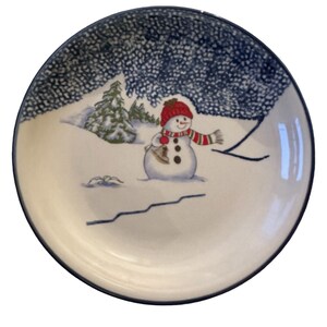 May include: A round, white ceramic plate with a winter scene. A snowman wearing a red hat and scarf holds a bell. The plate has a blue rim and a snowy forest background.