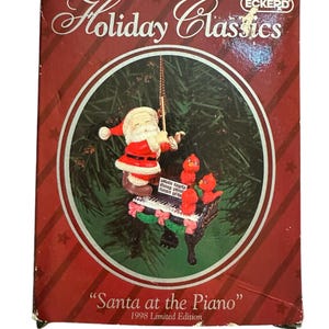 Puede incluir: Un adorno de Santa Claus rojo y blanco tocando un piano con dos cardenales rojos posados en la parte superior. El adorno está en una caja con las palabras "Holiday Classics" y "Santa at the Piano" 1998 Limited Edition.