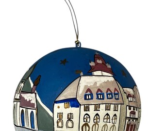 May include: A blue and white Christmas ornament depicting a snowy village scene with a church, houses, and a small boat on a frozen lake. The ornament has a gold string for hanging.