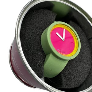 Pu&ograve; includere: Un orologio da polso verde e rosa con un bordo giallo attorno al quadrante. L'orologio &egrave; in una scatola di latta verde e argento con imbottitura in schiuma nera.