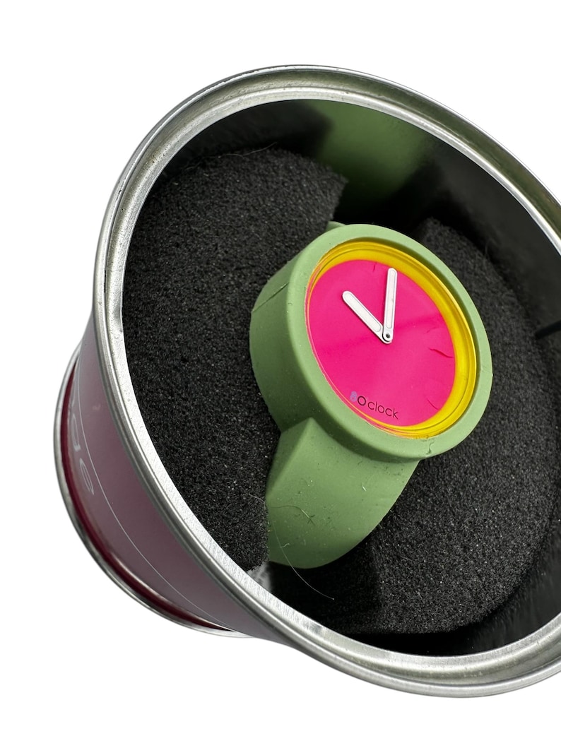 Pu&ograve; includere: Un orologio da polso verde e rosa con un bordo giallo attorno al quadrante. L'orologio &egrave; in una scatola di latta verde e argento con imbottitura in schiuma nera.