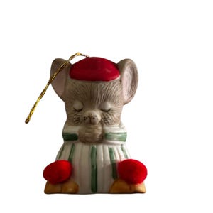 Puede incluir: Un adorno navideño de cerámica de un ratón con un gorro rojo y un suéter a rayas verdes y blancas. El ratón está sentado con las patas juntas y tiene un pompón rojo en cada pie.