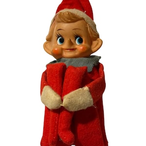 May include: A vintage elf figurine featuring a molded plastic face with painted blue eyes, rosy cheeks, and a pointed nose. The elf wears a red felt hat with a white trim, a grey felt collar, and a red felt body with white felt mittens.