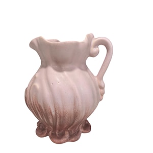 Puede incluir: Una jarra de cerámica rosa claro con borde y base con volantes. La jarra tiene un asa y un cuerpo bulboso con estrías verticales. La parte inferior de la jarra es de un tono marrón más oscuro.