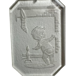 May include: A clear glass ornament with a raised design of a child standing near a fireplace. The child is wearing a long-sleeved shirt and pants. The fireplace has a brick chimney and a stocking hanging from it.