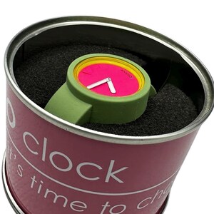 Pu&ograve; includere: Un orologio da polso rosa e verde con un bordo giallo attorno al quadrante. L'orologio &egrave; in un contenitore cilindrico rosa con le parole "It's time to clock" stampate sul lato.