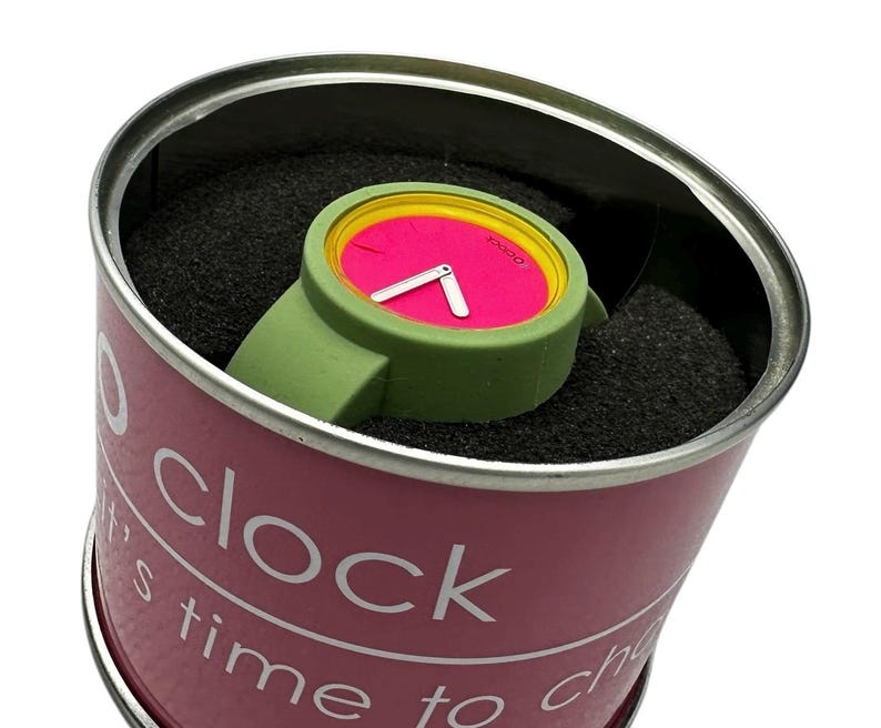 Pu&ograve; includere: Un orologio da polso rosa e verde con un bordo giallo attorno al quadrante. L'orologio &egrave; in un contenitore cilindrico rosa con le parole "It's time to clock" stampate sul lato.