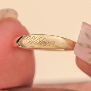 Può includere: Un anello d'oro con il nome "Wilson" inciso. L'anello è tenuto tra due dita. L'anello ha una superficie liscia e lucida con il nome inciso in corsivo.