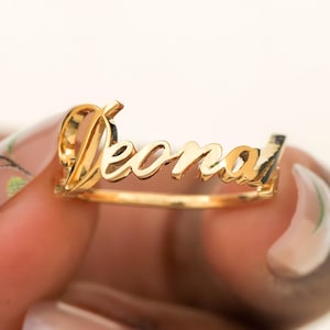 Puede incluir: Un anillo dorado con el nombre "Deonai" en fuente cursiva. El anillo se sostiene sobre un fondo claro. Es una pieza de joyería.