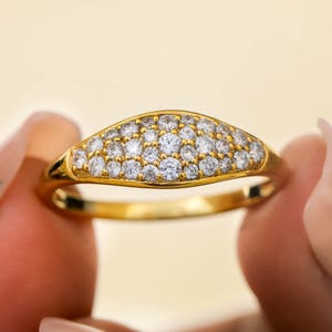 May include: A gold ring with a curved, oval-shaped top, set with numerous small, clear gemstones. The ring is held between two fingers against a soft yellow background, highlighting its polished, reflective surface.