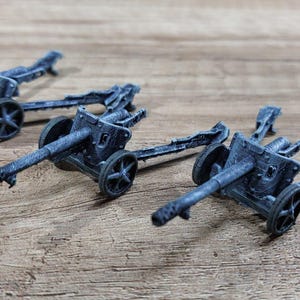 May include: Three miniature grey artillery cannons featuring long barrels, spoked wheels, and extended trail legs. These small-scale models are cast in a matte grey finish, showcasing detailed mechanical components including the breech and carriage.